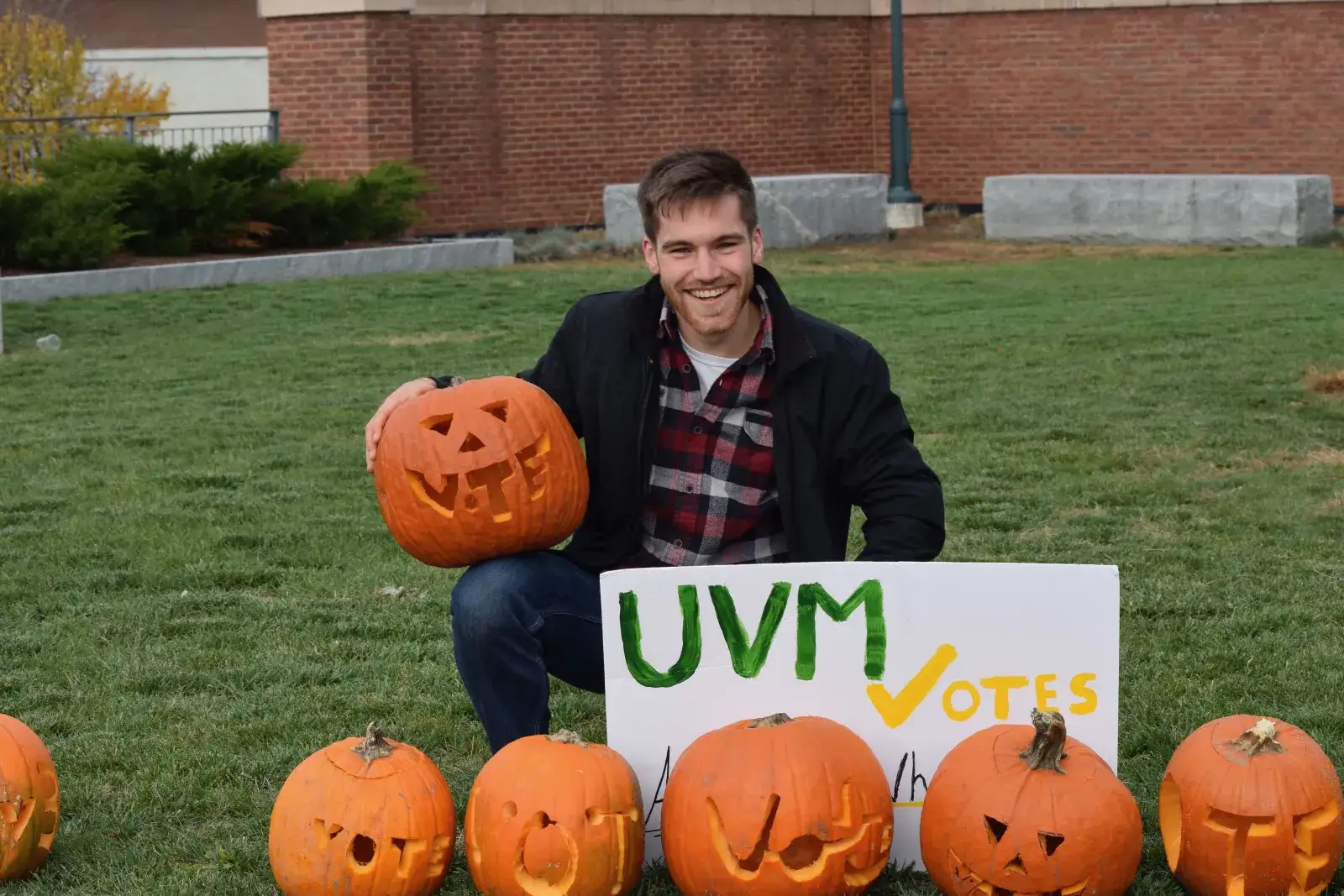 A student kneeling on a campus lawn behind a row of jack-o-lanterns carved with the word VOTE, next to a hand-painted UVM Votes sign