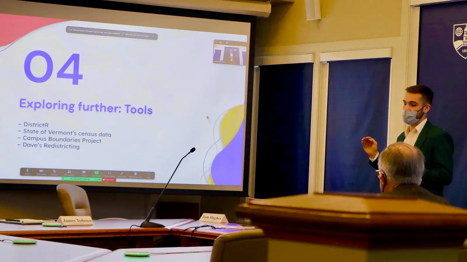 Presenting redistricting tools to a seated audience in a wood-paneled meeting room, projector screen showing a slide titled "Exploring further: Tools"