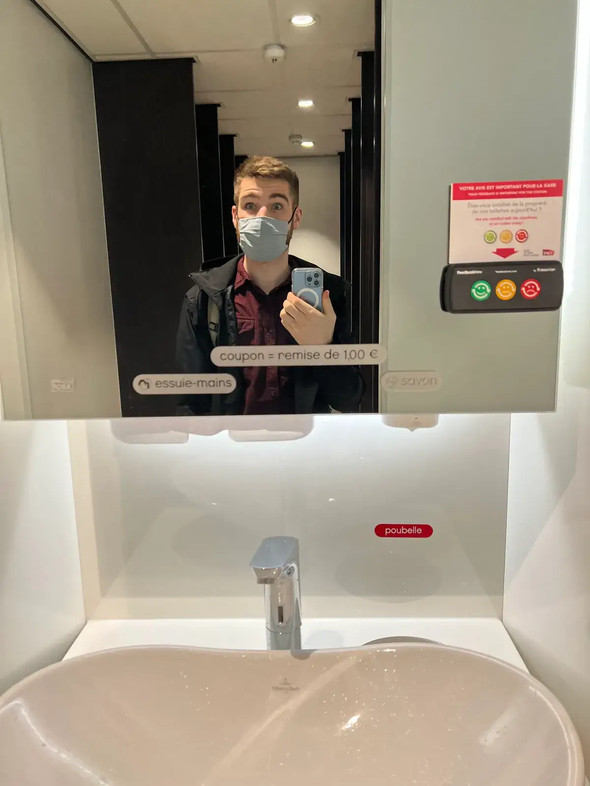 Mirror selfie in an SNCF station restroom, where every fixture is labeled — essuie-mains, savon, poubelle — beside a smiley-button cleanliness feedback panel